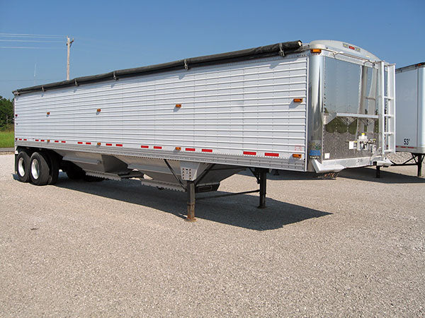 Side view of a Timpte aluminum grain trailer parked on a gravel lot, featuring a ribbed aluminum body and a roll tarp "Kwik-Lock" system along the top, shown under clear daylight conditions.