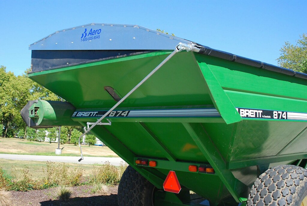 Green Brent 874 grain cart with large tires and an attached blue Aero cover, parked outdoors on a sunny day with trees and grass in the background.