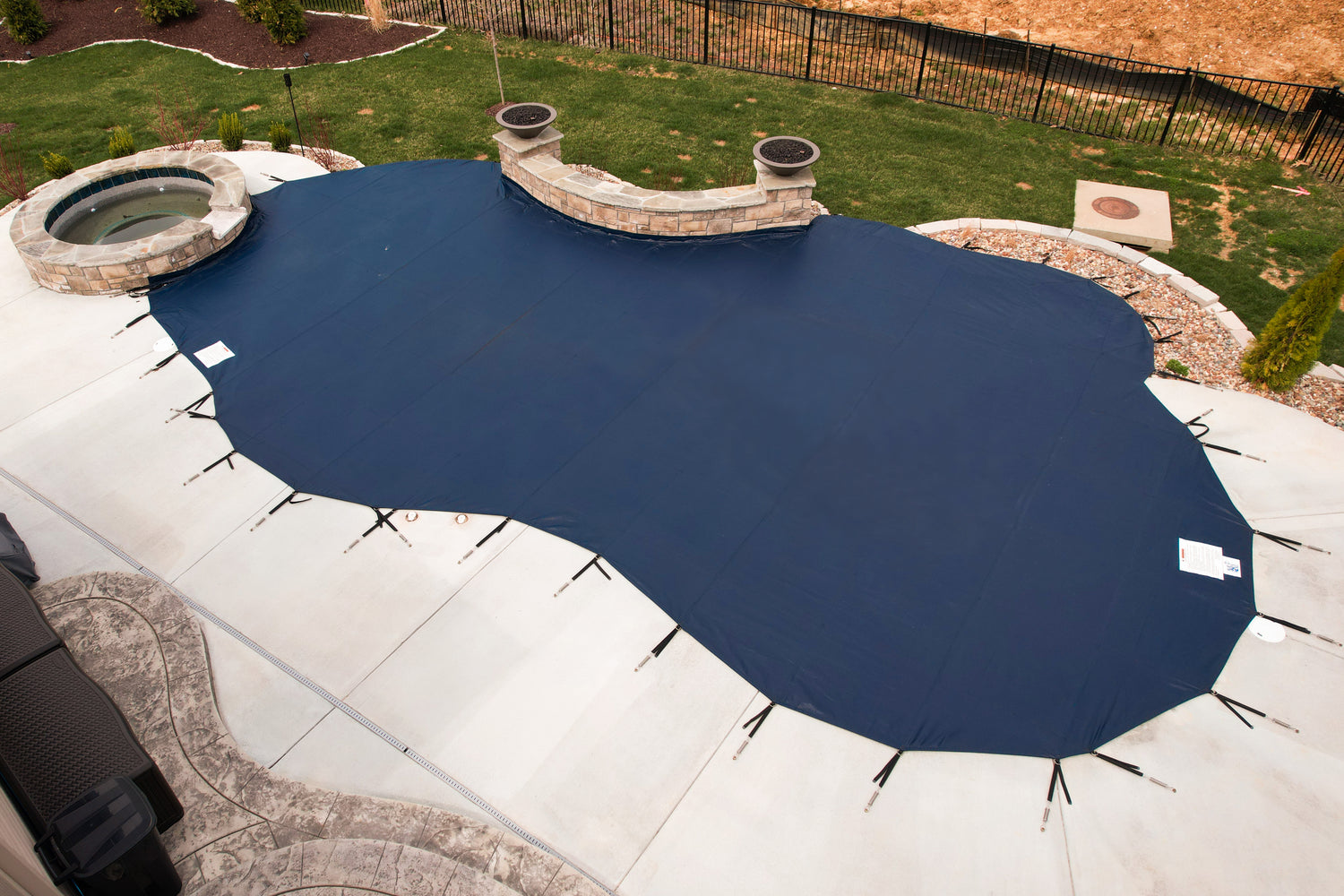 Dark blue mesh safety cover secured with straps over a freeform in-ground swimming pool, adjacent to a raised circular spa and a curved stone retaining wall, with a lawn, fence, and landscaped yard in the background.