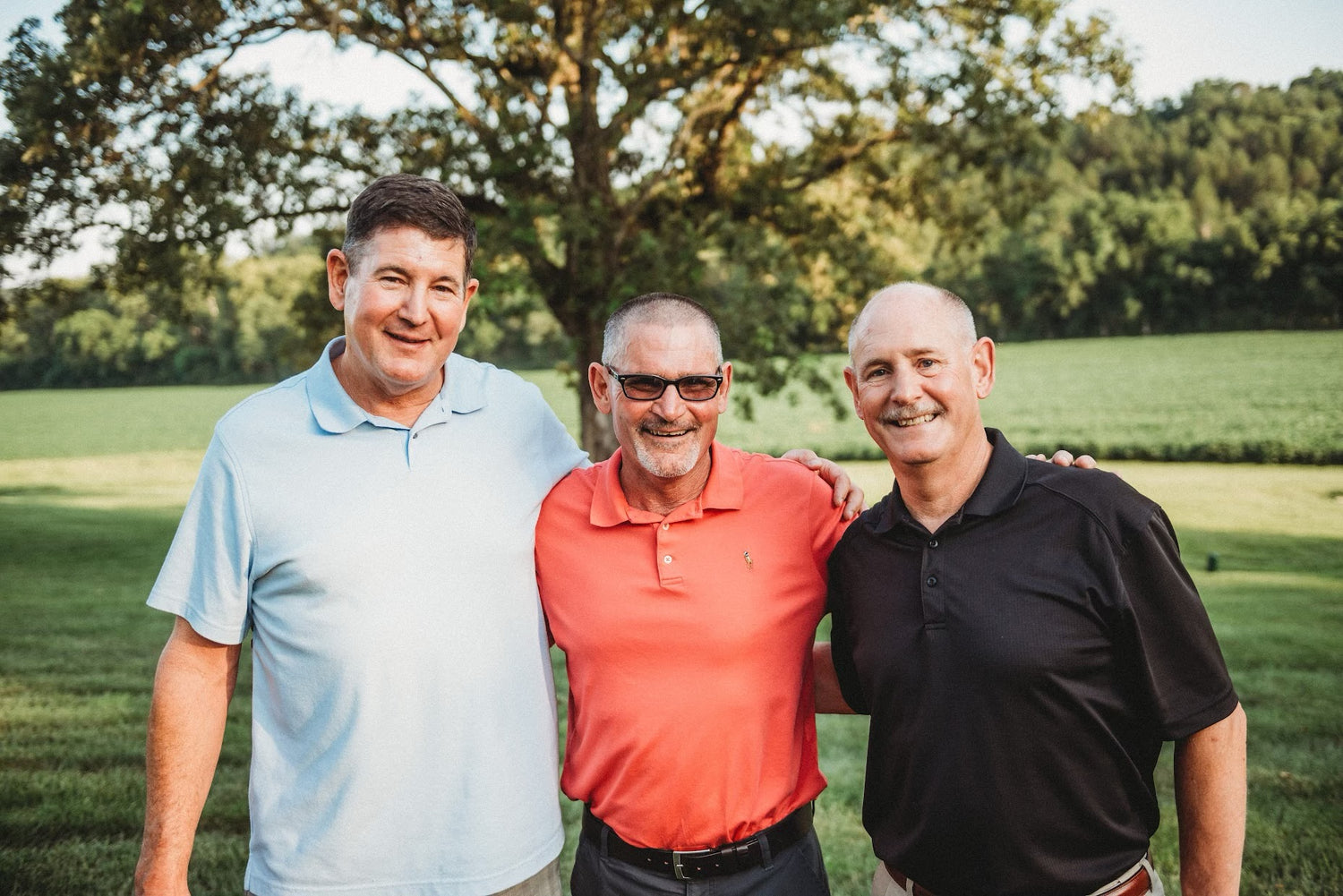 Three men stand together outdoors on a sunny day, smiling at the camera. The man on the left wears a light blue polo shirt, the man in the center wears a red-orange polo shirt and glasses, and the man on the right wears a black polo shirt. They have their arms around each other, with a large tree and green field in the background.