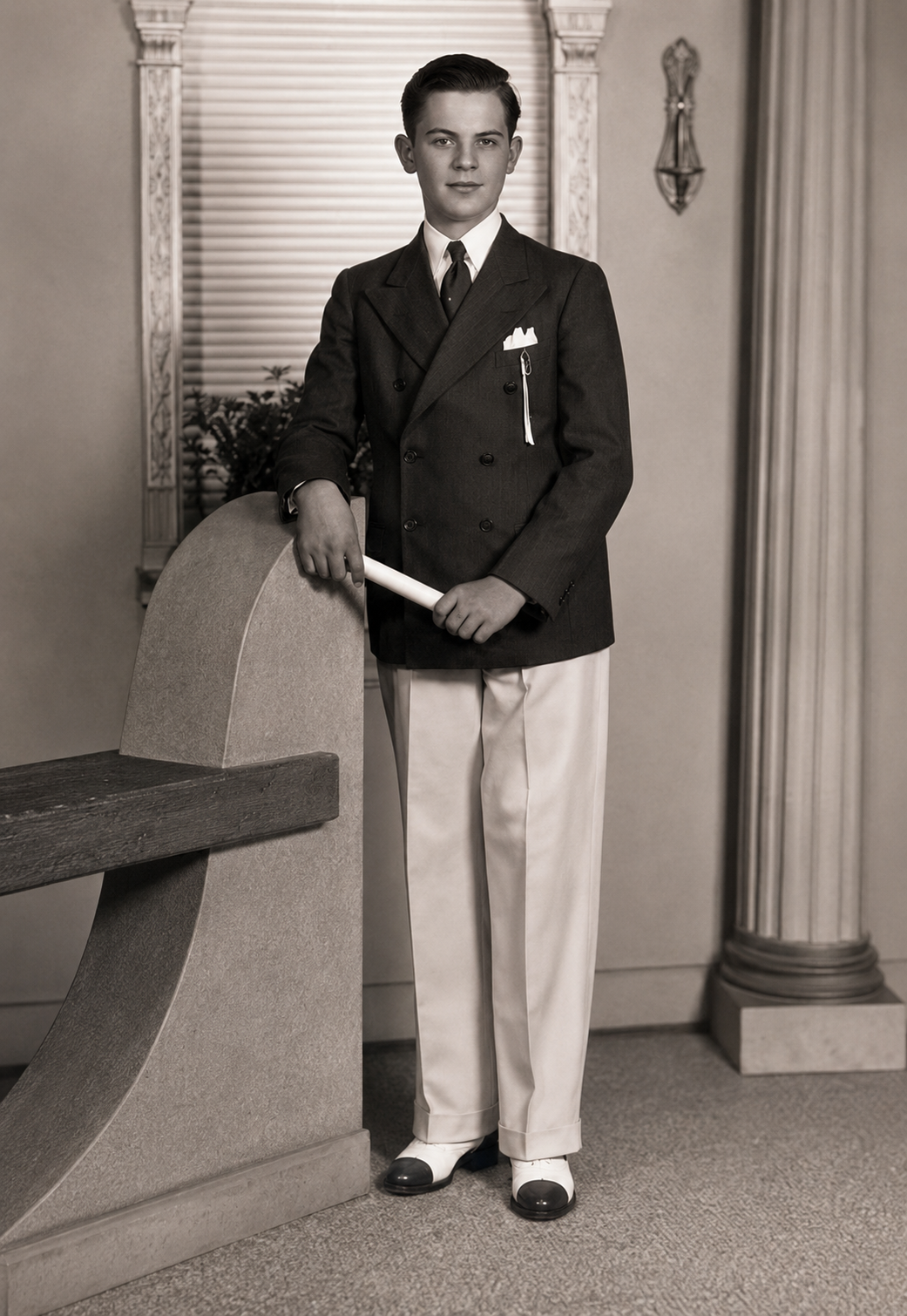 A black and white vintage portrait of a young boy standing in a formal suit, holding a rolled diploma or scroll, posed indoors next to a stone bench with columns behind him.