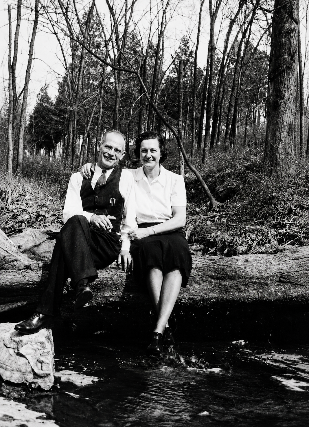 Historic black-and-white photograph of two adults seated on a fallen log beside a shallow stream in a wooded area, with leafless trees and dense forest vegetation in the background.