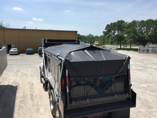 Rear view of a dump truck equipped with a Kwik‑Lock roll tarp system, showing a black tarp covering the load and secured along the sides, parked in an industrial yard with buildings and trees in the background.
