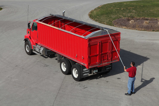 Red dump truck with an open-top bed and partially rolled tarp cover being operated with a manual crank, parked on a paved surface.